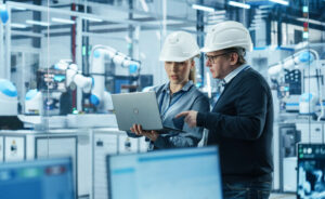 Male Engineer and Female Project Manager Standing Next to an Assembly Line, Talking, Using Computer and Monitoring Production at a Modern Automated Electronics Manufacture with AI Robotic Arms